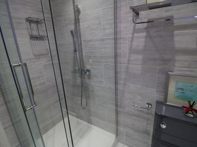 A shower area with glass doors grey tiled walls a hand shower a towel rack and a small cabinet with a framed picture and a diffuser at Flat 3 Peacehaven Sandsend near Whitby