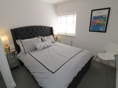 A bedroom with a double bed lamps bedside tables a window a framed picture on the wall and a white chair at Flat 3 Peacehaven Sandsend near Whitby