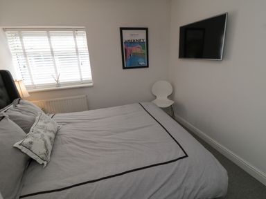 A bedroom with a bed grey bedding a wall-mounted television a white chair and a window with blinds at Flat 3 Peacehaven in Sandsend near Whitby