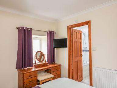 A bedroom with a wooden dressing table and mirror purple curtains and a door leading to a bathroom at Sorrel Cottage in Padstow