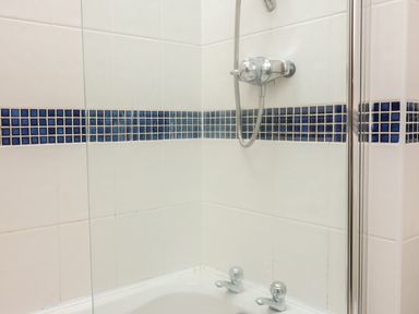 A bathroom corner with white tiles and blue mosaic border with a shower head and taps at Sorrel Cottage in Padstow