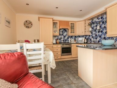 A kitchen with wooden cabinets blue tiled backsplash a dining table with white chairs and a red sofa at Sorrel Cottage in Padstow