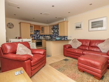 A living room with red leather sofas and a kitchen with wooden cabinets and blue tiled backsplash at Sorrel Cottage in Padstow