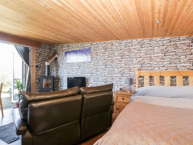 A bedroom with a bed a wooden nightstand a black leather sofa a wood stove and stone patterned wallpaper at Blackbrae Cabin in Glenboig near Coatbridge Glasgow and Stirling