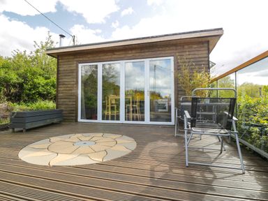 A wooden deck with a circular stone pattern in front of a cabin with glass doors and metal chairs at Blackbrae Cabin in Glenboig near Coatbridge Glasgow and Stirling