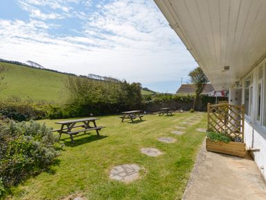 A garden with picnic tables next to a white building and green fields at 2 Brook Cottages in Hope Cove