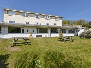 A building with white walls and multiple windows with picnic tables on a grassy area in front at 2 Brook Cottages in Hope Cove