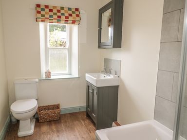 A bathroom with a toilet a window with a checkered blind a sink with a cabinet a basket on the floor and a bathtub at Newbiggin Cottage in Blanchland