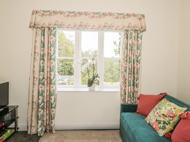 A window with floral curtains and a white orchid on the sill next to a teal sofa with colorful cushions at Newbiggin Cottage in Blanchland