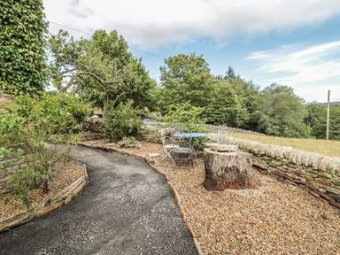 A garden path with gravel and a tree stump table with metal chairs surrounded by stone walls and trees at Newbiggin Cottage in Blanchland