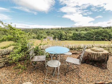An outdoor seating area with metal chairs and a round table on a gravel surface near a stone wall overlooking a field and trees at Newbiggin Cottage in Blanchland
