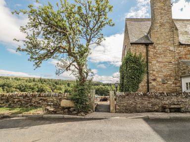 A stone wall with an open gate and a tree next to a stone house with ivy on the wall at Newbiggin Cottage in Blanchland