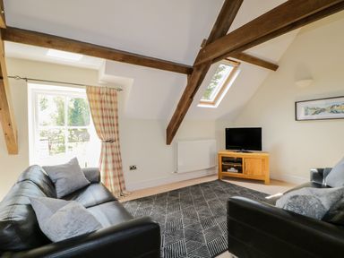A living room with two black leather sofas a patterned rug a wooden TV stand with a television exposed wooden beams and a window with checkered curtains at Carpenters Cottage in Cockermouth