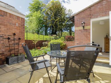 An outdoor patio area with a glass table and four black chairs next to a brick building with a garden and trees in the background at Carpenters Cottage in Cockermouth