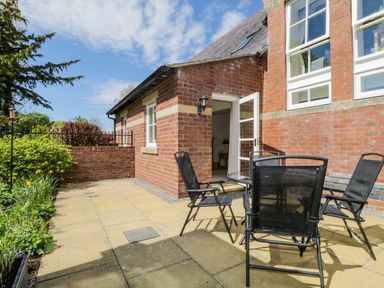 A patio with a round glass table and black chairs next to a red brick building with an open door at Carpenters Cottage in Cockermouth
