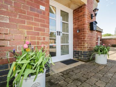 The entrance of a brick building with white double doors two flower pots with pink and purple flowers on a paved ground at Carpenters Cottage in Cockermouth