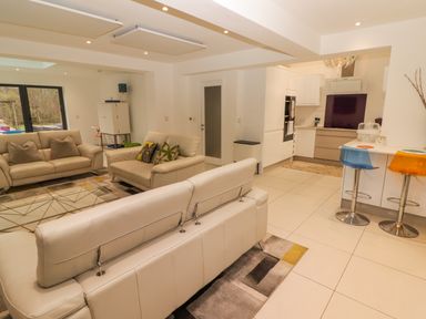 A living room with beige sofas and a glass coffee table next to a kitchen with a breakfast bar and colorful stools at Hiraeth in Cwmtaf near Cefn-Coed-Y-Cymmer