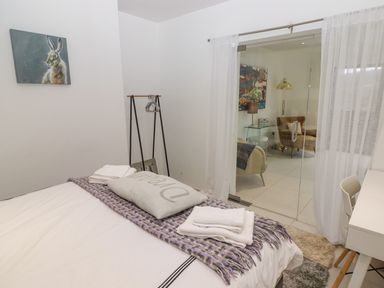 A bedroom with a bed covered in white sheets and a purple throw blanket a pillow with text a small clothing rack and a glass door opening to a sitting area at Hiraeth in Cwmtaf near Cefn-Coed-Y-Cymmer