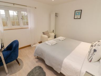 A bedroom with a bed covered with a gray quilt and pillows a white armchair a blue chair by a window with sheer curtains and a small rug on the floor at Hiraeth in Cwmtaf near Cefn-Coed-Y-Cymmer