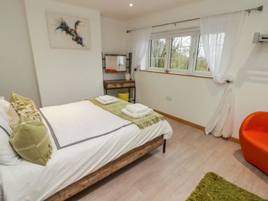 A bedroom with a bed with pillows towels and a green throw a window with white curtains a wooden vanity and an orange chair at Hiraeth in Cwmtaf near Cefn-Coed-Y-Cymmer