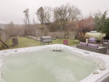 An outdoor hot tub with water flowing near a barbecue grill and patio furniture in a fenced backyard at Hiraeth in Cwmtaf near Cefn-Coed-Y-Cymmer