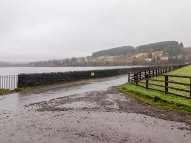 A wet road next to a stone wall and wooden fence near a lake with hills and trees in the background at Hiraeth in Cwmtaf near Cefn-Coed-Y-Cymmer