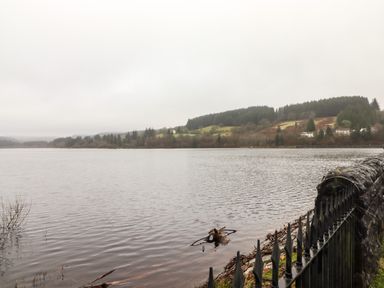 A lake with wooded hills and houses in the background and a black iron fence in the foreground at Hiraeth in Cwmtaf near Cefn-Coed-Y-Cymmer