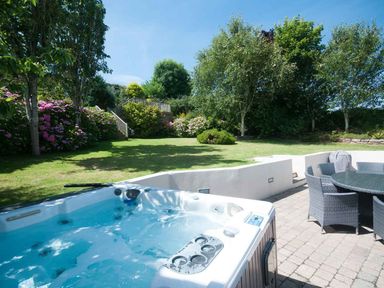 An outdoor patio with a hot tub and a round table with chairs beside a grassy garden with trees and flowers at The Orchard in Crantock