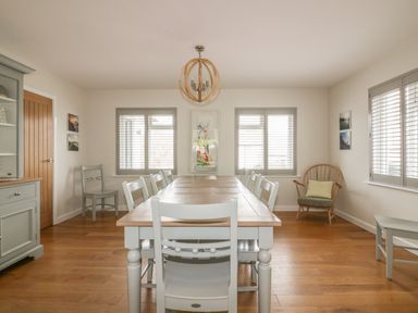 A dining room with a long wooden table and chairs with windows and a wooden cabinet at The Orchard in Crantock