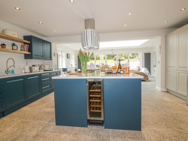 A kitchen with blue cabinets a central island with a wine cooler and glass doors leading to a garden at The Orchard in Crantock
