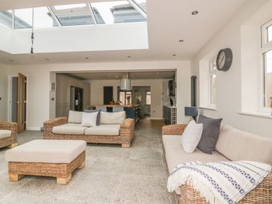 A living room with wicker sofas and cushions a coffee table and a kitchen area with island and chairs at The Orchard in Crantock
