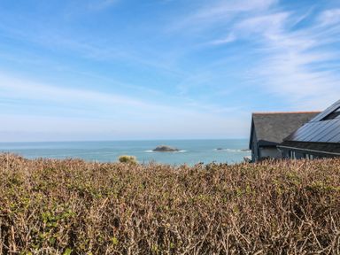 A view of the ocean with rocks and a hedge in the foreground and a house with solar panels on the roof