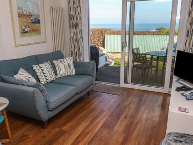 A living room with a blue sofa and geometric patterned cushions next to sliding glass doors opening to a garden and sea view at Kittiwake in Crantock