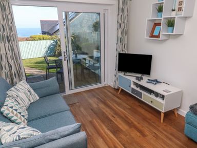 A living room with a blue sofa a TV on a white stand and sliding glass doors leading to a garden at Kittiwake in Crantock