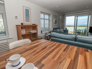 A living room with a wooden table, blue sofas, bookshelf, window blinds, and sliding glass doors leading to an outdoor seating area at Kittiwake in Crantock