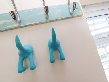 Two blue dog tail-shaped wall hooks on a white wall beneath a coat rack with chrome hooks and a window with white blinds