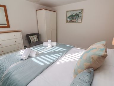 A bedroom with a bed covered with light blue bedding and rolled towels a white wardrobe a chest of drawers a chair and a framed picture on the wall at Kittiwake in Crantock