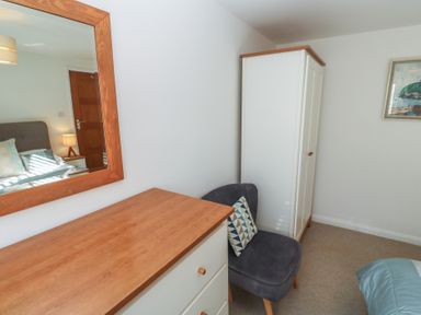 A bedroom corner with a white wardrobe a blue chair with a patterned cushion a wooden dresser and a wooden framed mirror reflecting a bed and door at Kittiwake in Crantock