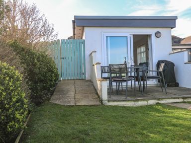 A patio with a table and four chairs outside a building with glass sliding doors and a lawn with bushes at Kittiwake Crantock