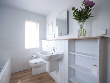 A bathroom with a sink, toilet, bathtub, shelves with a roll of toilet paper, and flowers in a vase on the counter at Kareena in Crantock