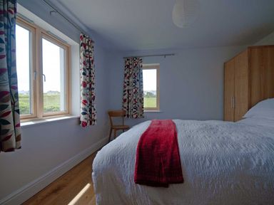 A bedroom with a bed covered in white linens and a red throw blanket two windows with patterned curtains a wooden chair and a wooden wardrobe at Kareena in Crantock
