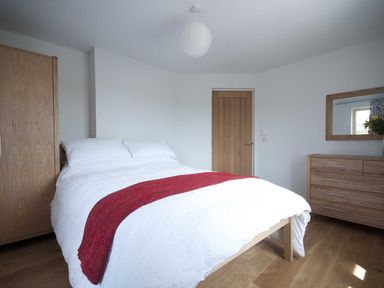 A bedroom with a bed white bedding and red runner wooden wardrobe wooden door and wooden dresser with mirror at Kareena in Crantock