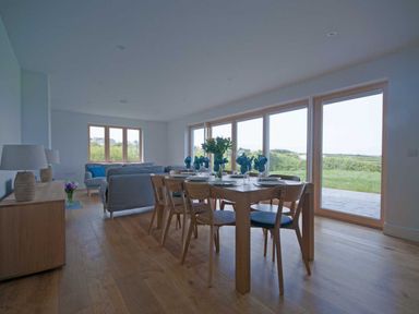 A dining table set with blue napkins and plates beside sofas and large windows showing greenery at Kareena in Crantock