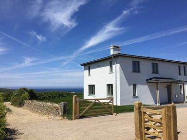 A white two-story house with multiple windows and wooden gates on a gravel driveway with a stone wall and greenery nearby and a blue sky in the background at Kareena in Crantock