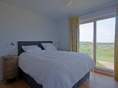 A bedroom with a double bed white bedding wooden nightstands and yellow floral curtains with a glass door overlooking green fields at Kareena in Crantock
