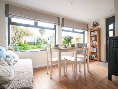 A dining area with a white table and chairs a cushioned bench bookshelf and large windows showing a garden at Keelyn in Crantock