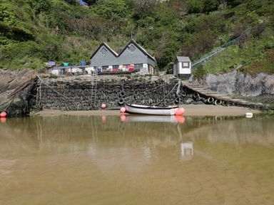 A small boat moored at a stone dock with a ramp and buildings on a hillside at Keelyn in Crantock