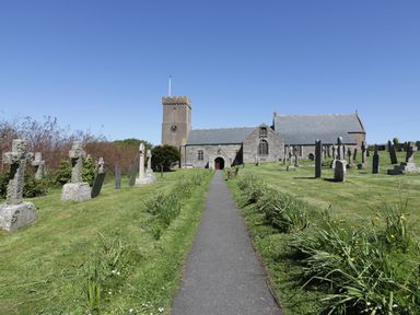 A pathway leading to a stone church surrounded by a graveyard with tombstones and grass at Keelyn in Crantock