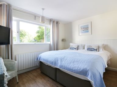 A bedroom with a large bed with blue and white bedding a window with curtains a wicker chair a wall-mounted television and a framed picture at Keelyn in Crantock