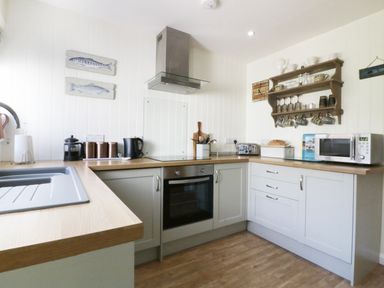A kitchen with white cabinets a wooden countertop a sink microwave oven toaster and wall shelves with cups at Keelyn in Crantock
